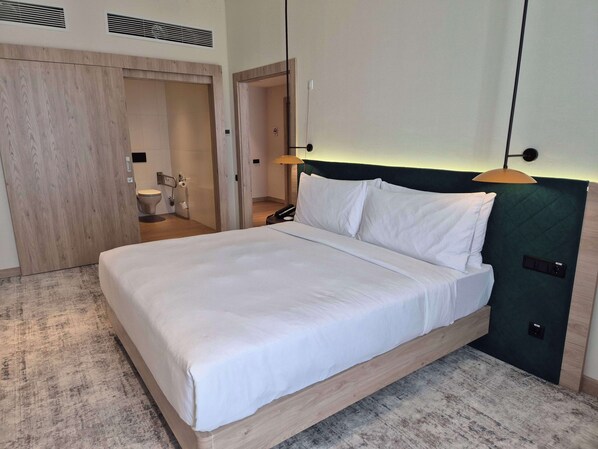 A modern hotel room with a wooden bed frame, white linens, a green headboard, and a visible bathroom with a toilet and sink. - Hilton Garden Inn Termez Airitom (Termez)