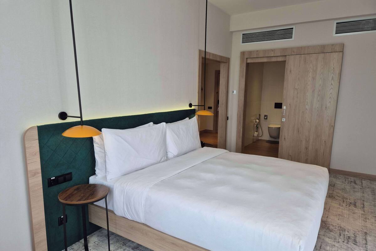 Une chambre d’hôtel moderne avec un grand lit, une petite table et une salle de bain visible à travers une porte ouverte.