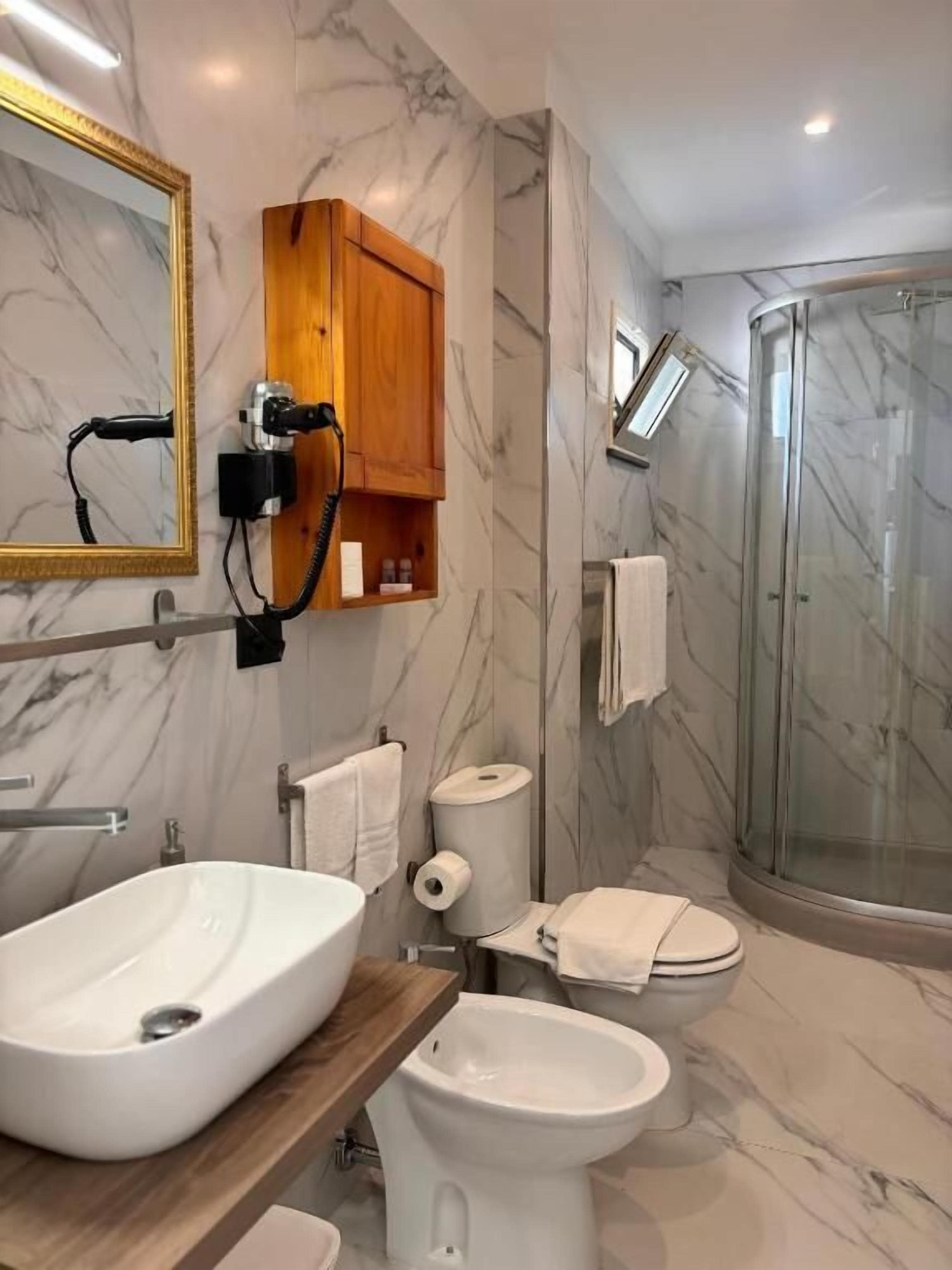 Une salle de bain avec un lavabo en marbre, des toilettes, un bidet, une douche et un meuble en bois contenant un sèche-cheveux.