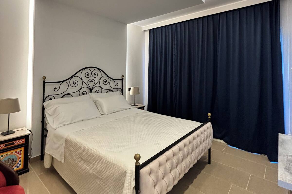 Ein Schlafzimmer mit einem Bett, einem Nachttisch, einer Lampe, einem roten Sessel und einem großen Fenster mit dunklen Vorhängen.