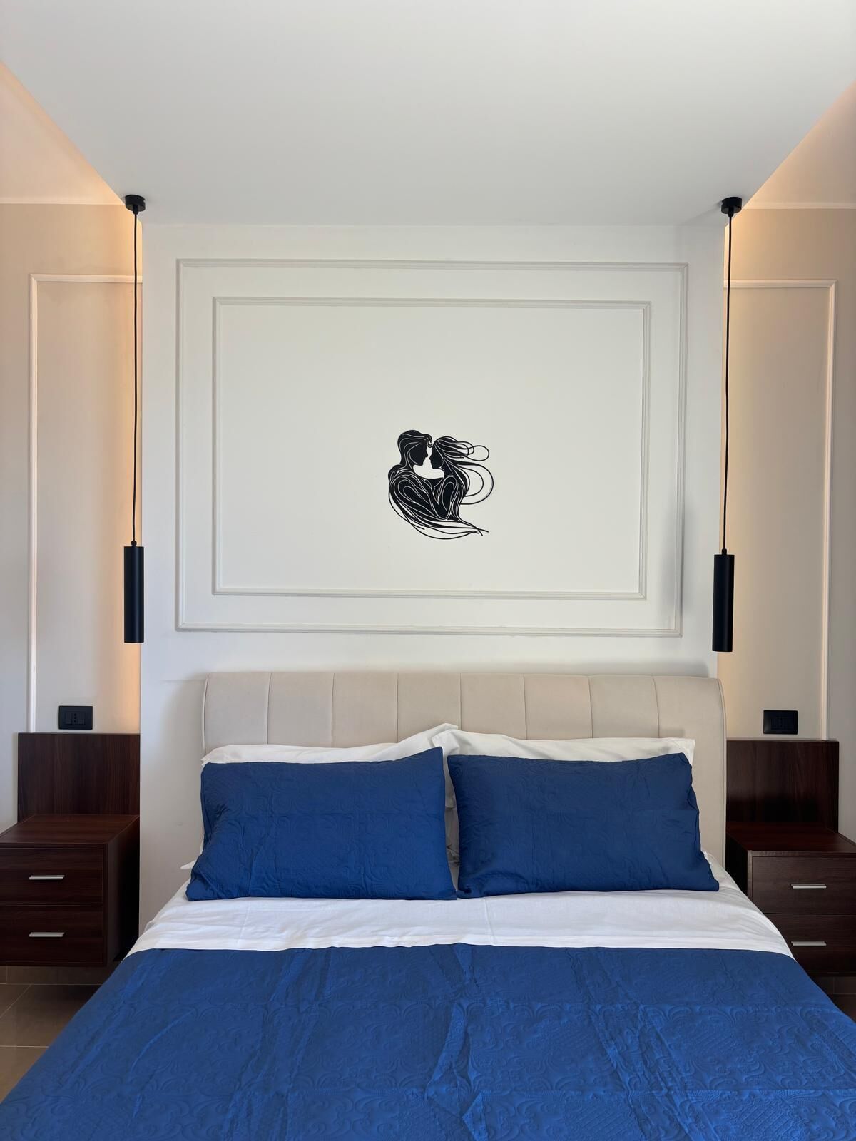 A bedroom with a bed, two nightstands, a wall-mounted artwork, and a ceiling light fixture.