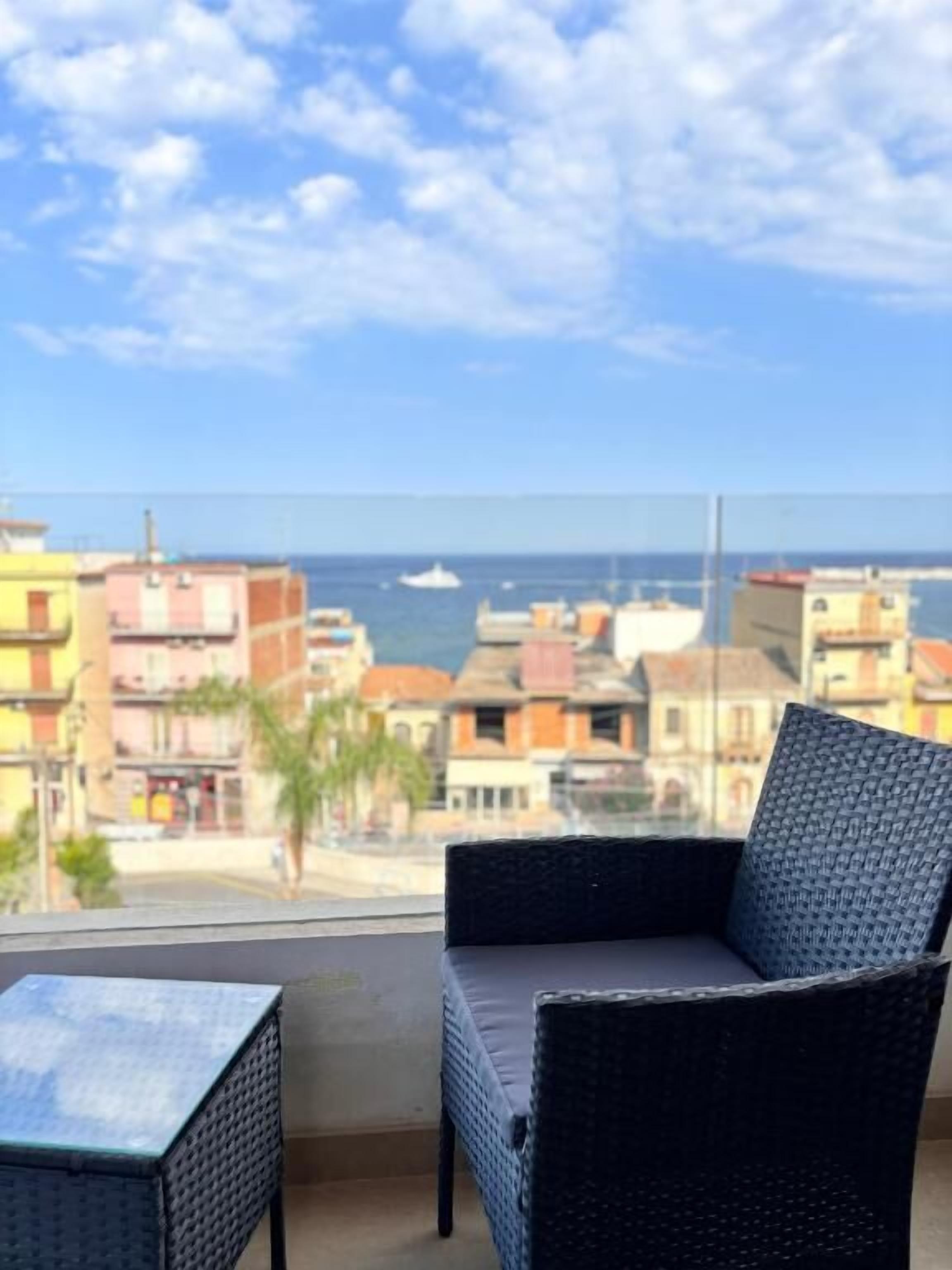 A balcony with a wicker chair and a small table, overlooking a coastal cityscape with buildings and the sea.
