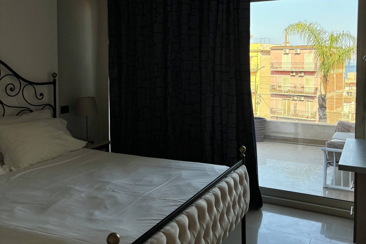 Ein Schlafzimmer mit Bett, einem Fenster mit Blick auf Gebäude und einem Balkon.
