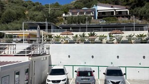 A parking area with three cars, a building with multiple balconies, and a hill with vegetation in the background. - IG - Villa Don Antonio  Apartment  (Giardini Naxos)