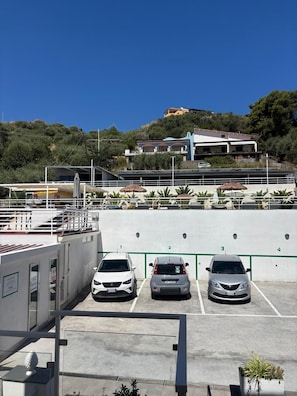 A parking area with three cars, a building with multiple balconies, and a hill with vegetation in the background. - IG - Villa Don Antonio  Apartment  (Giardini Naxos)