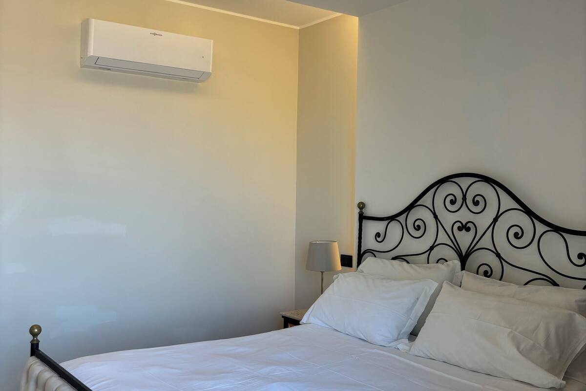 Ein ordentlich bezogenes Bett mit Kopfteil, einer Nachttischlampe und einer Klimaanlage an der Wand.