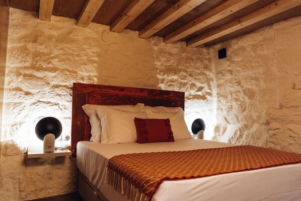 A bedroom with a wooden headboard, a bed with white bedding and a red pillow, two wall-mounted lamps, and a stone wall.