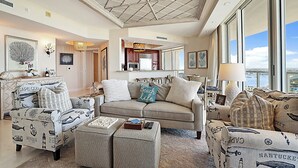 A spacious living room with nautical-themed furniture, a large window offering a city view, and a modern kitchen in the background. - Charming 3-bedroom condo  (West Palm Beach)