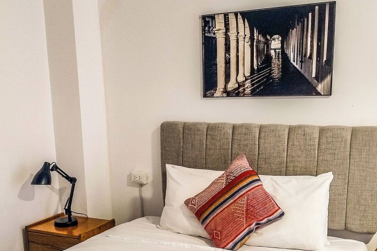 A neatly made bed with a patterned blanket, a pillow with a colorful design, a wall-mounted picture, and a bedside lamp.