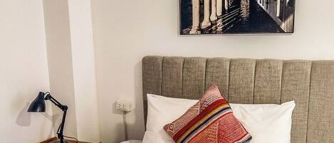 A neatly made bed with a patterned blanket, a pillow with a colorful design, a wall-mounted picture, and a bedside lamp.