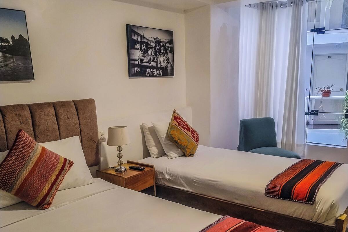 A hotel room with two beds, a nightstand with a lamp, a chair, and a window with curtains.