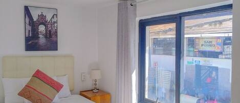 A hotel room with a bed, a nightstand, a lamp, a window with a view of the street, and a table with a chair.