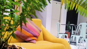 A yellow sofa with a colorful striped pillow, a wicker basket, and a potted plant in a modern outdoor setting with tiled flooring and light-colored walls. - Central Market Hotel San Pedro (Cusco)
