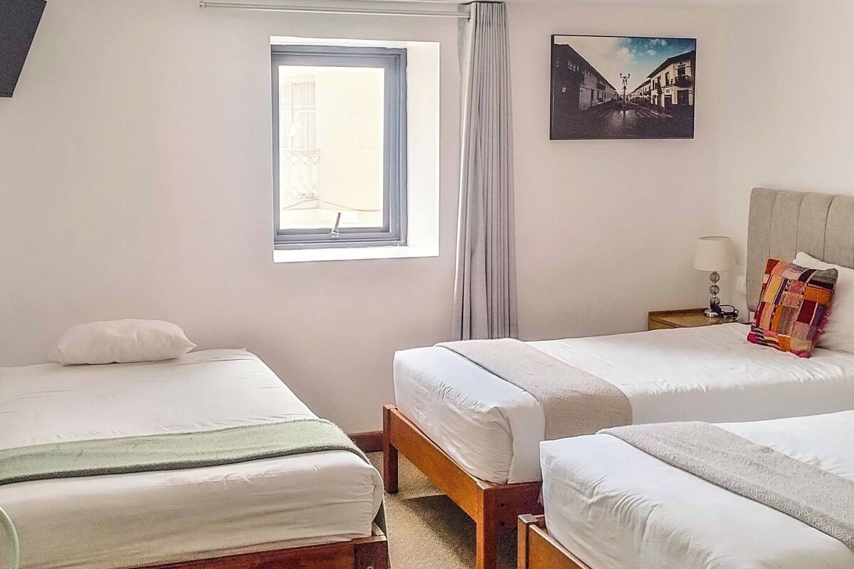 A hotel room with two single beds, a window with curtains, a painting on the wall, and a bedside table with a lamp.