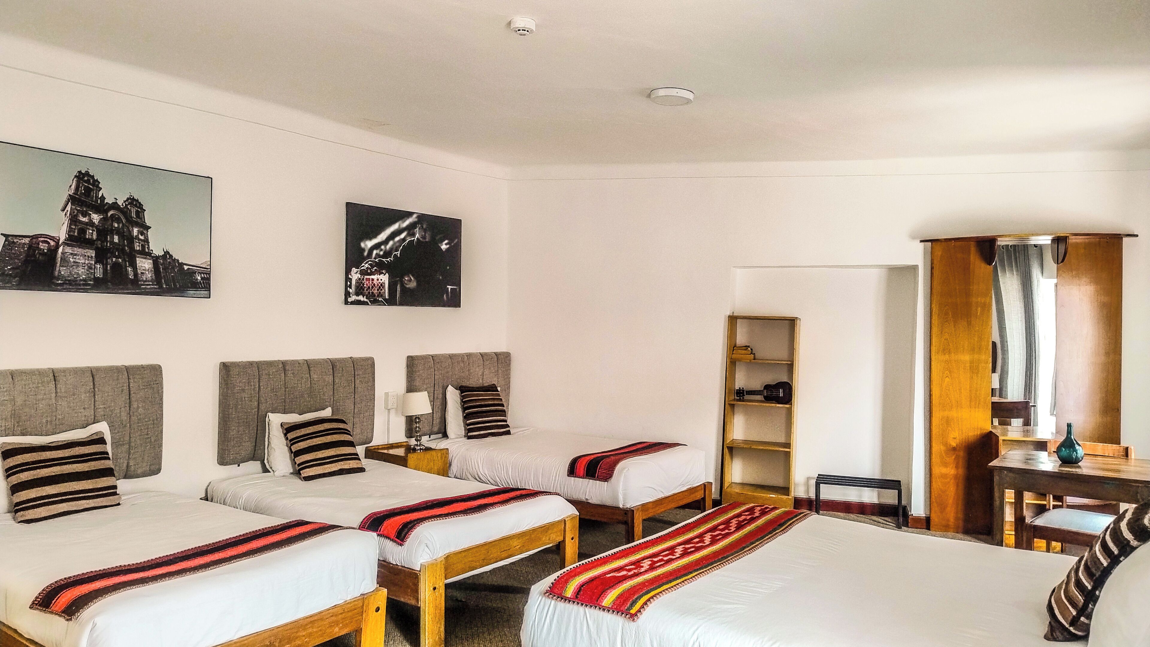 Habitación de hotel con dos camas, un escritorio de madera, una estantería y dos cuadros colgados en la pared.