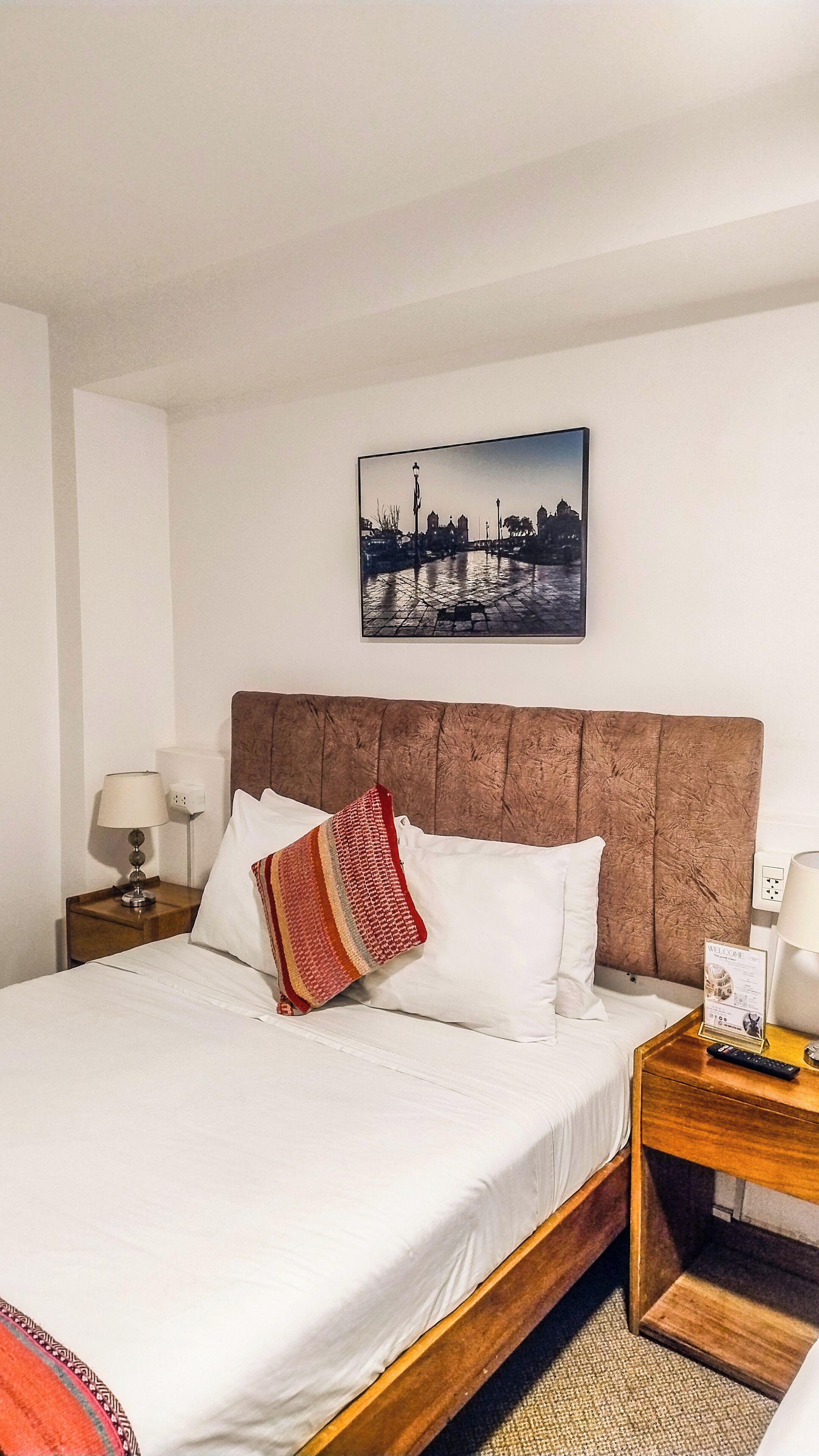 Ein Hotelzimmer mit einem Bett, einem Nachttisch, einer Lampe, einem Willkommensschild und einem Bild an der Wand.