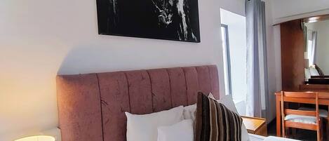 Habitación de hotel con una cama grande, cabecero, mesita de noche con lámpara, una silla de madera y un escritorio con silla.