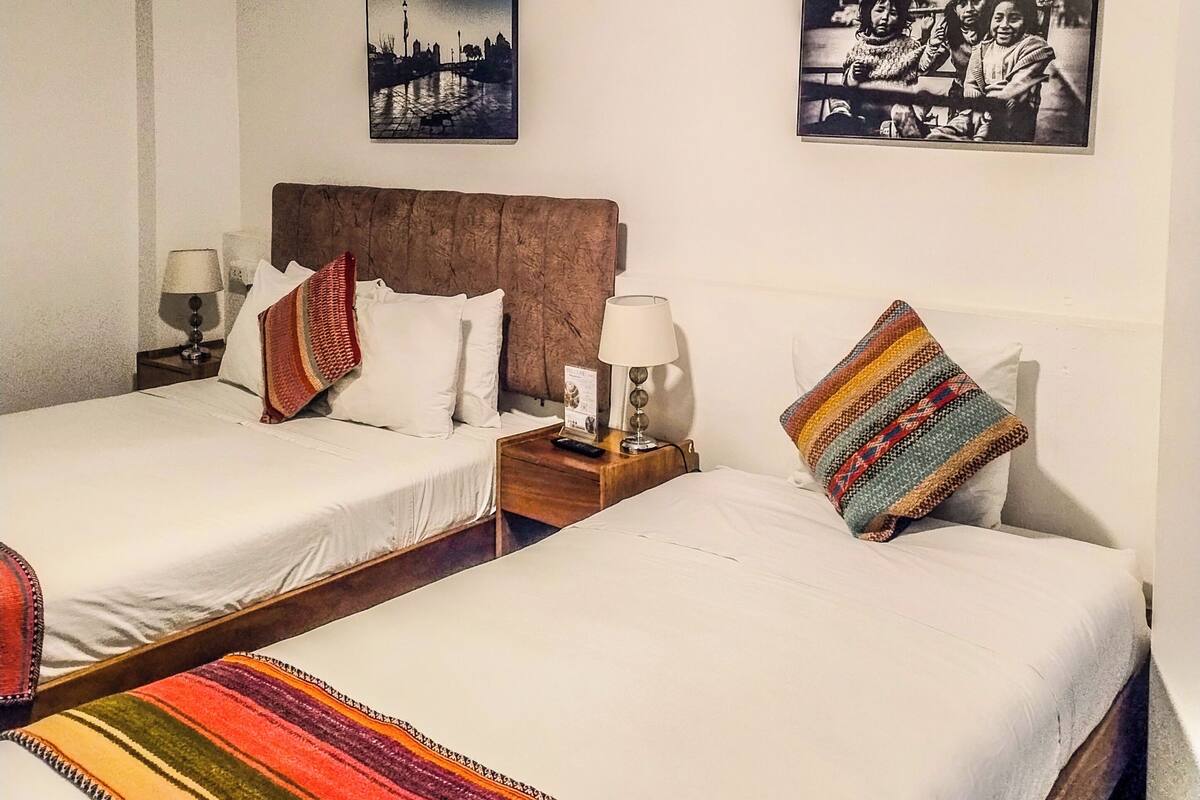 A hotel room with two beds, a wooden headboard, a nightstand with a lamp, and two wall-mounted pictures.