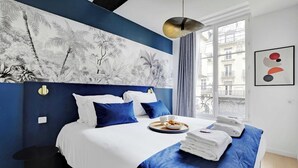 Una camera d'albergo moderna con un grande letto, biancheria azzurra e bianca, un murale tropicale, una finestra con vista sugli edifici e un quadro astratto incorniciato.