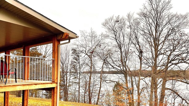 A wooden deck with a railing, overlooking a grassy area with trees and a body of water in the background.
