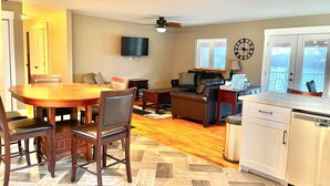 A spacious living area with a wooden dining table, comfortable seating, a ceiling fan, and a kitchen with a dishwasher.