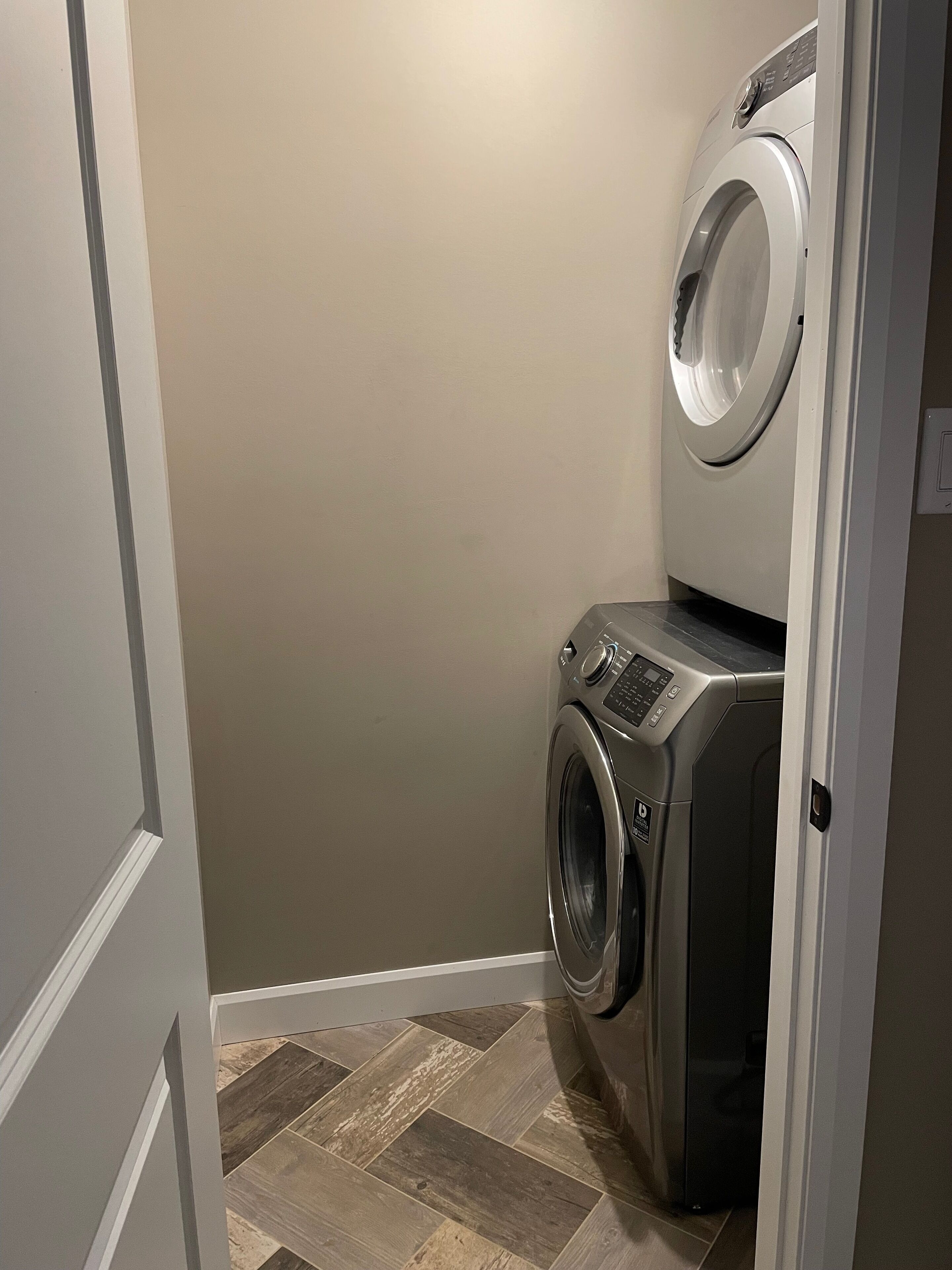 A laundry room with a front-loading washer and dryer, wooden herringbone floor, and a door leading to another room.