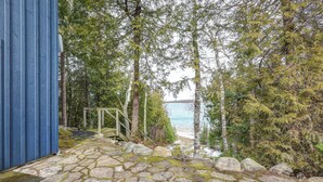 A stone pathway leading to a wooden deck with a view of a lake and forest. - Cozy cottage nestled along 90ft of pristine Georgian Bay pebble beachfront (Wiarton)
