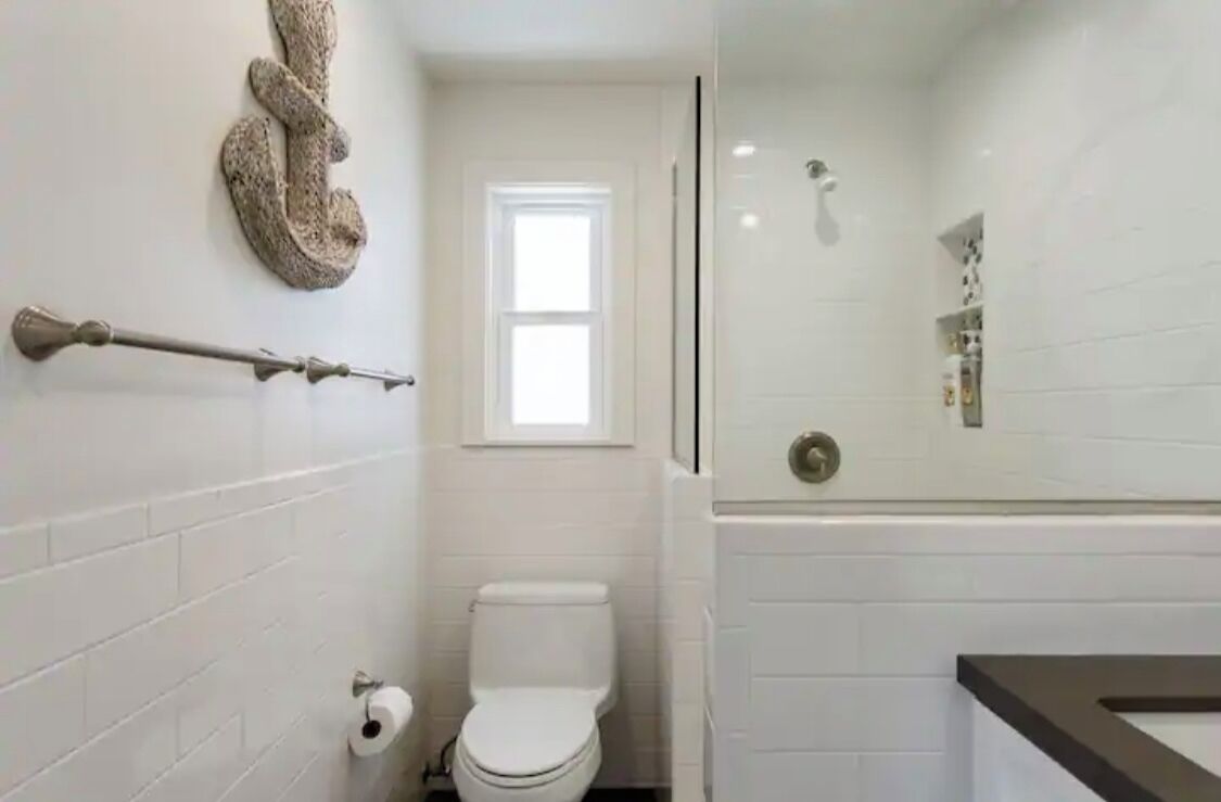 Un baño con paredes de azulejos blancos, un inodoro blanco, un perchero, una ducha con puerta de vidrio y un ventanal que deja entrar luz natural.