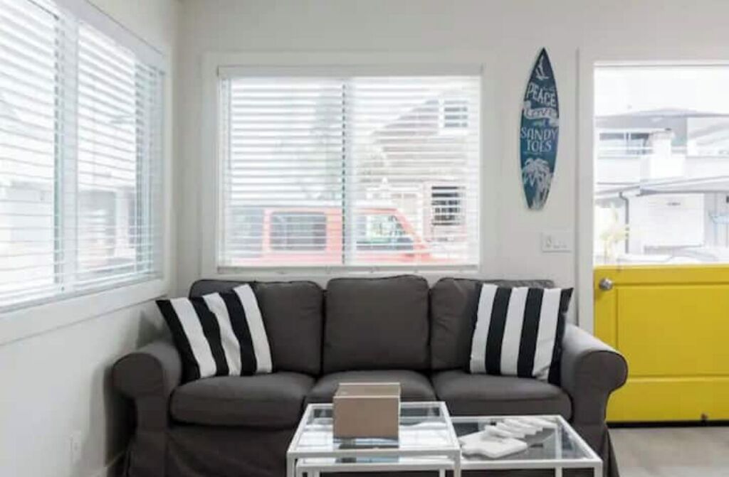 A living room with a grey sofa, striped pillows, a glass coffee table with a laptop, and a surfboard sign on the wall.