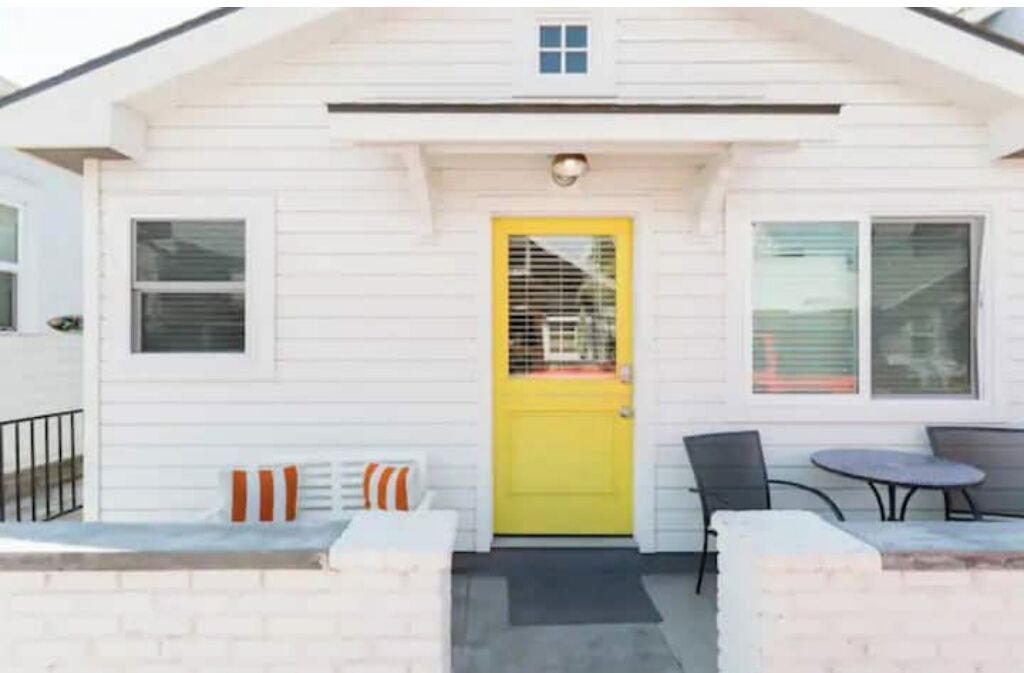 A white building with a yellow door, a small porch with a table and chairs, and a window with blinds.