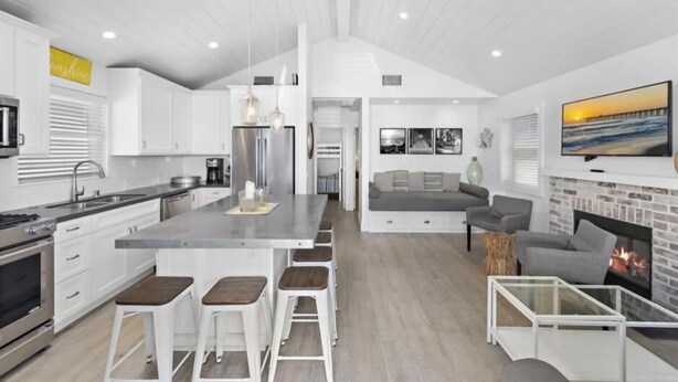 A modern kitchen with a central island, bar stools, and a dining area with a sofa and fireplace. - Good Day Sunshine Cottage-Newly updated!! Light and Airy w parking  (Newport Beach)