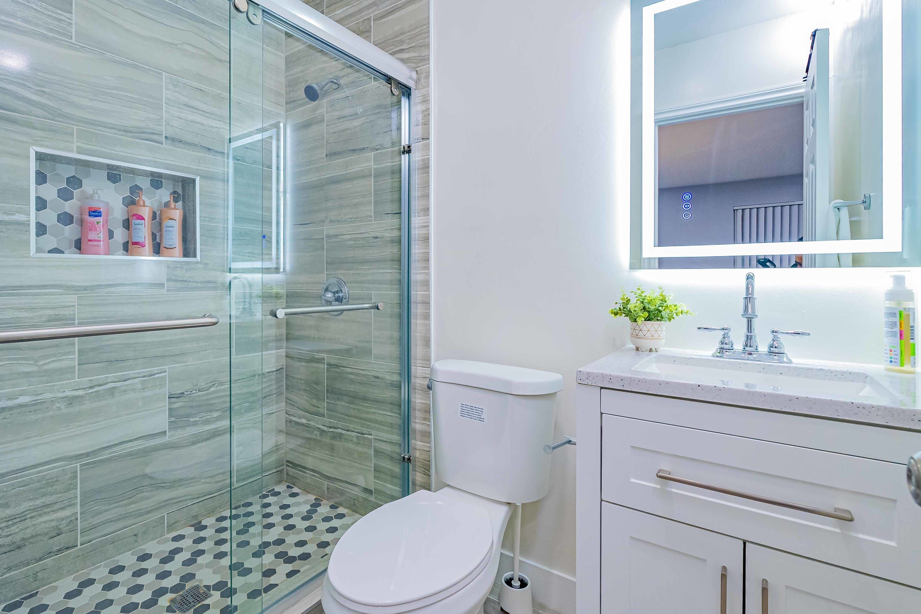 A modern bathroom with a glass-enclosed shower, a toilet, a sink with a mirror above it, and toiletries on the countertop.