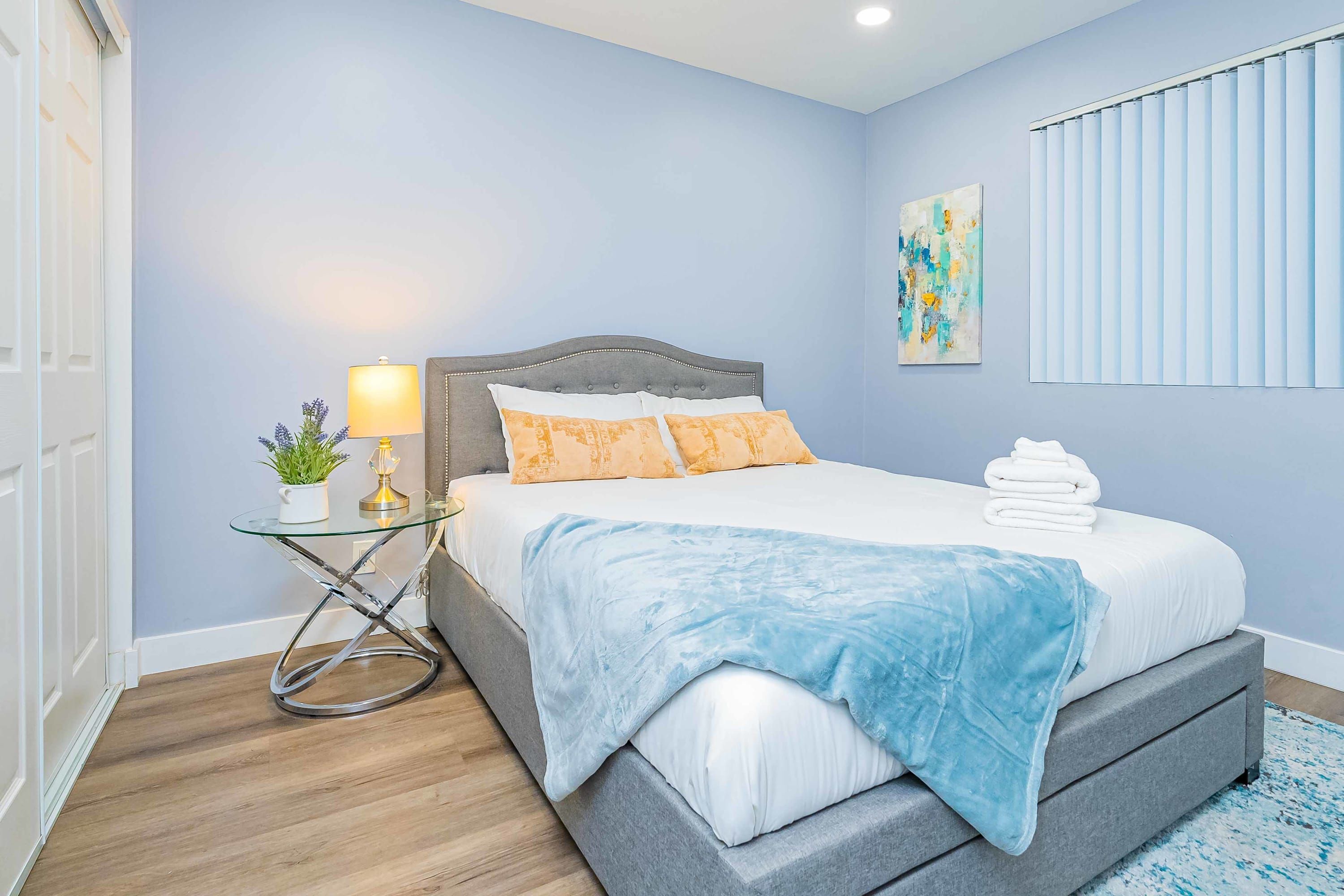 A bedroom with a bed, a bedside table with a lamp, a wall art piece, and a window with blinds.