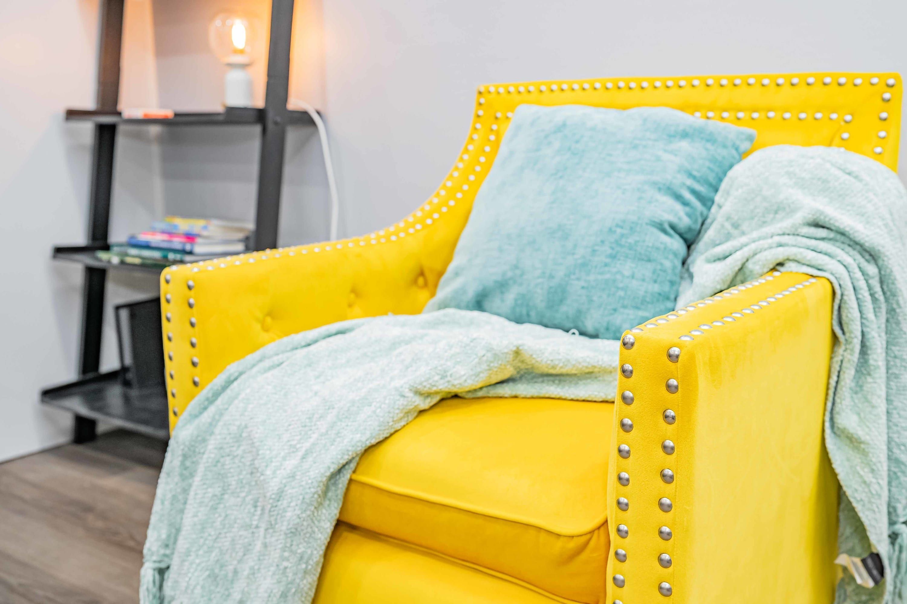 A yellow tufted armchair with a grey throw blanket, a blue pillow, and a metal studded frame.