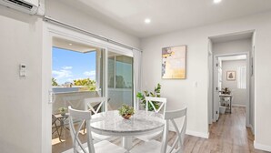 Habitación moderna y luminosa con una mesa de comedor de mármol y sillas blancas, una puerta corrediza de vidrio que da a un balcón, y un pasillo con una planta y obras de arte enmarcadas.