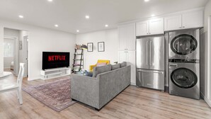 Una sala de estar moderna con un sofá gris, un televisor de pantalla plana mostrando 'NETFLIX', una lavadora y un refrigerador.