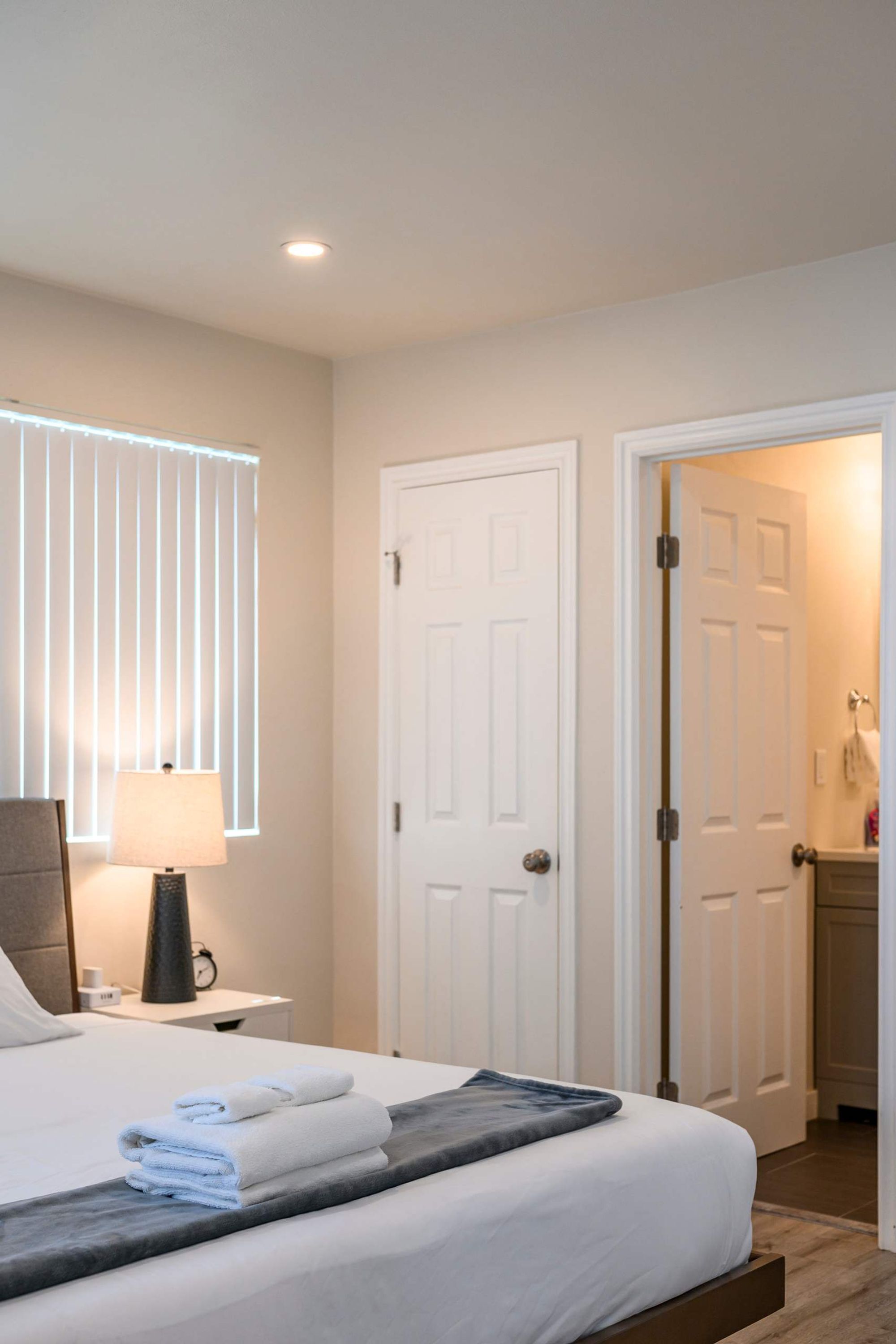 A hotel room with a neatly made bed, a bedside lamp, and a window with blinds.