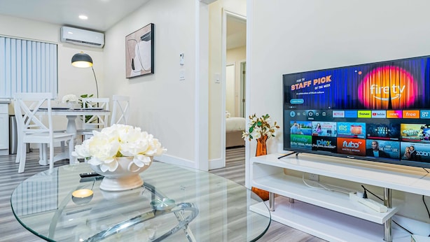 A modern living room with a glass-top coffee table, white furniture, a flat-screen TV displaying various streaming services, and a vase with white flowers. - Charming Elegant Retreat in the San Gabriel Valley (Altadena)