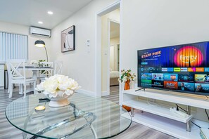 Ein modernes Wohnzimmer mit einem Glastisch, weißen Möbeln, einem Flachbildfernseher, der verschiedene Streaming-Dienste anzeigt, und einer Vase mit weißen Blumen.