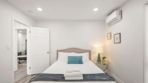 A neatly made bed with white linens and a blue pillow, a bedside table with a lamp, and an open door leading to a bathroom. - Charming Elegant Retreat in the San Gabriel Valley (Altadena)