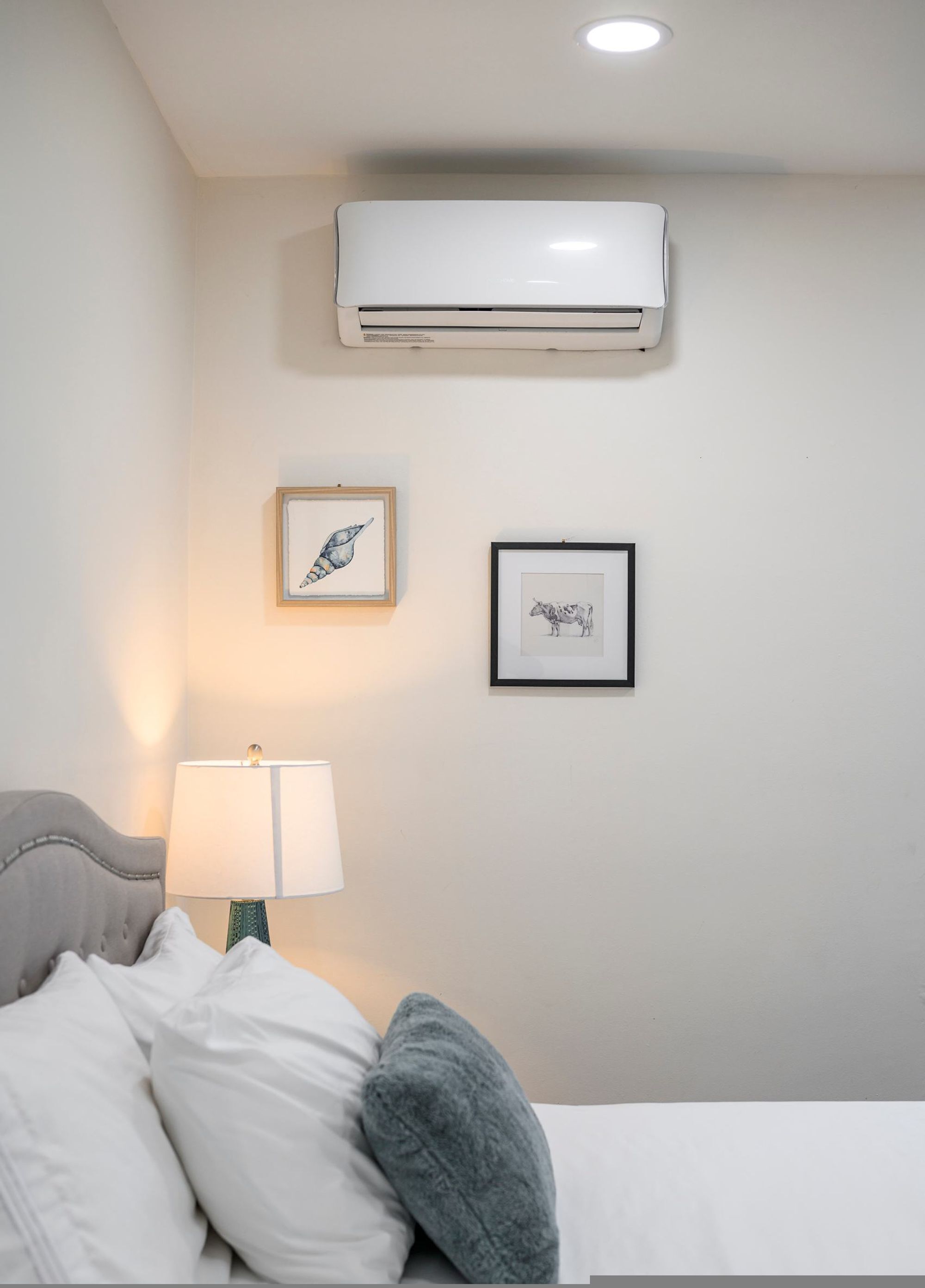 Ein Schlafzimmer mit einem Bett, einer Nachttischlampe, einer Klimaanlage und zwei gerahmten Bildern an der Wand.