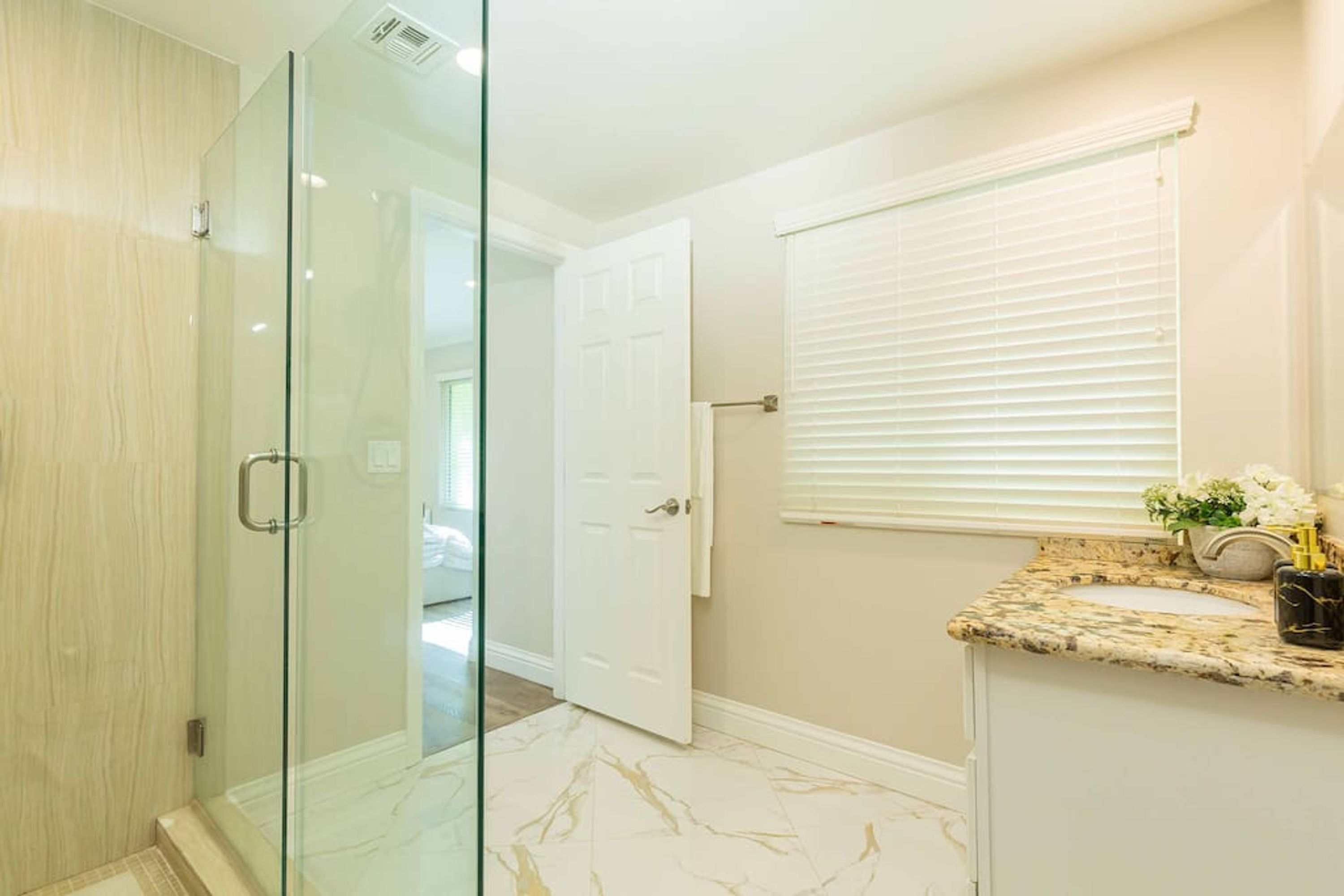 A bathroom with a glass shower enclosure, a white door, a window with blinds, and a marble-tiled floor.