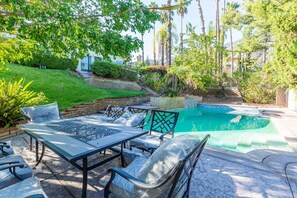 An outdoor pool area with a table and chairs, surrounded by greenery and palm trees.