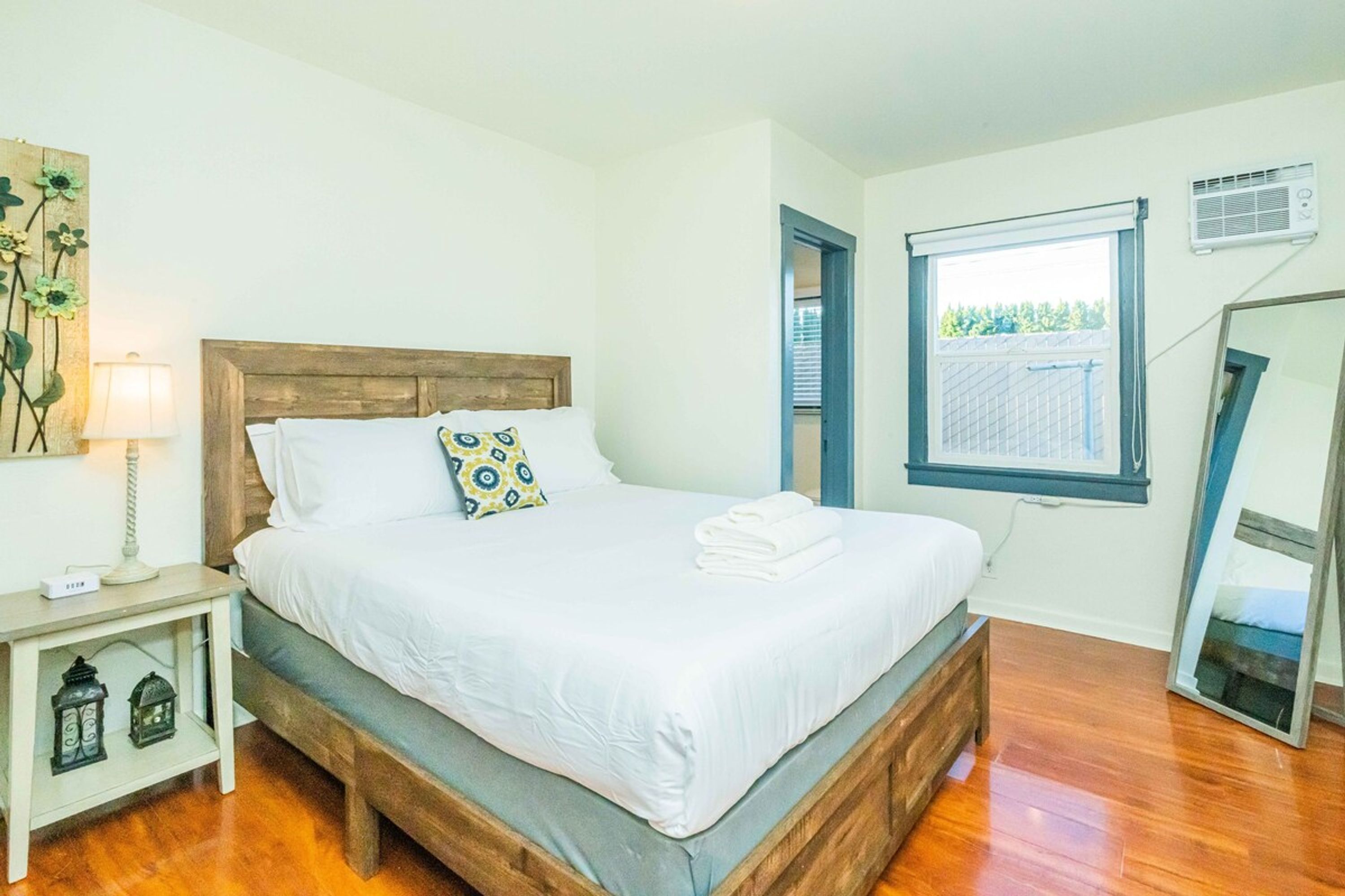Un dormitorio con una cama de madera, sábanas blancas, una mesita de noche con lámpara y una ventana que da a zonas verdes.