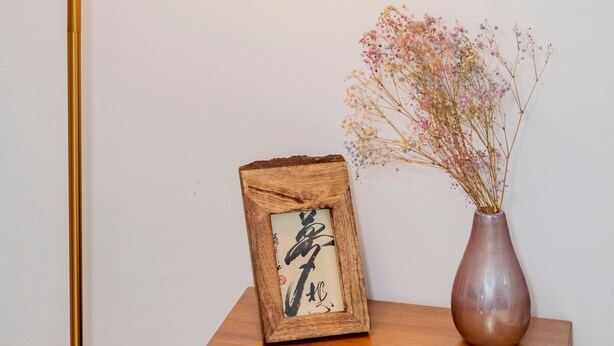 Ett träbord som står vid sängen, med en inramad bild och en vas med torkade blommor.