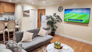 Un salon avec un canapé gris, une table basse, un téléviseur diffusant un événement sportif, un coin cuisine avec des meubles en bois et une porte.