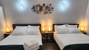 Une chambre d’hôtel avec deux lits, chacun recouvert de draps blancs et d’un plaid vert foncé. Il y a des tables de chevet avec des lampes, et un luminaire fixé au mur au-dessus des lits.