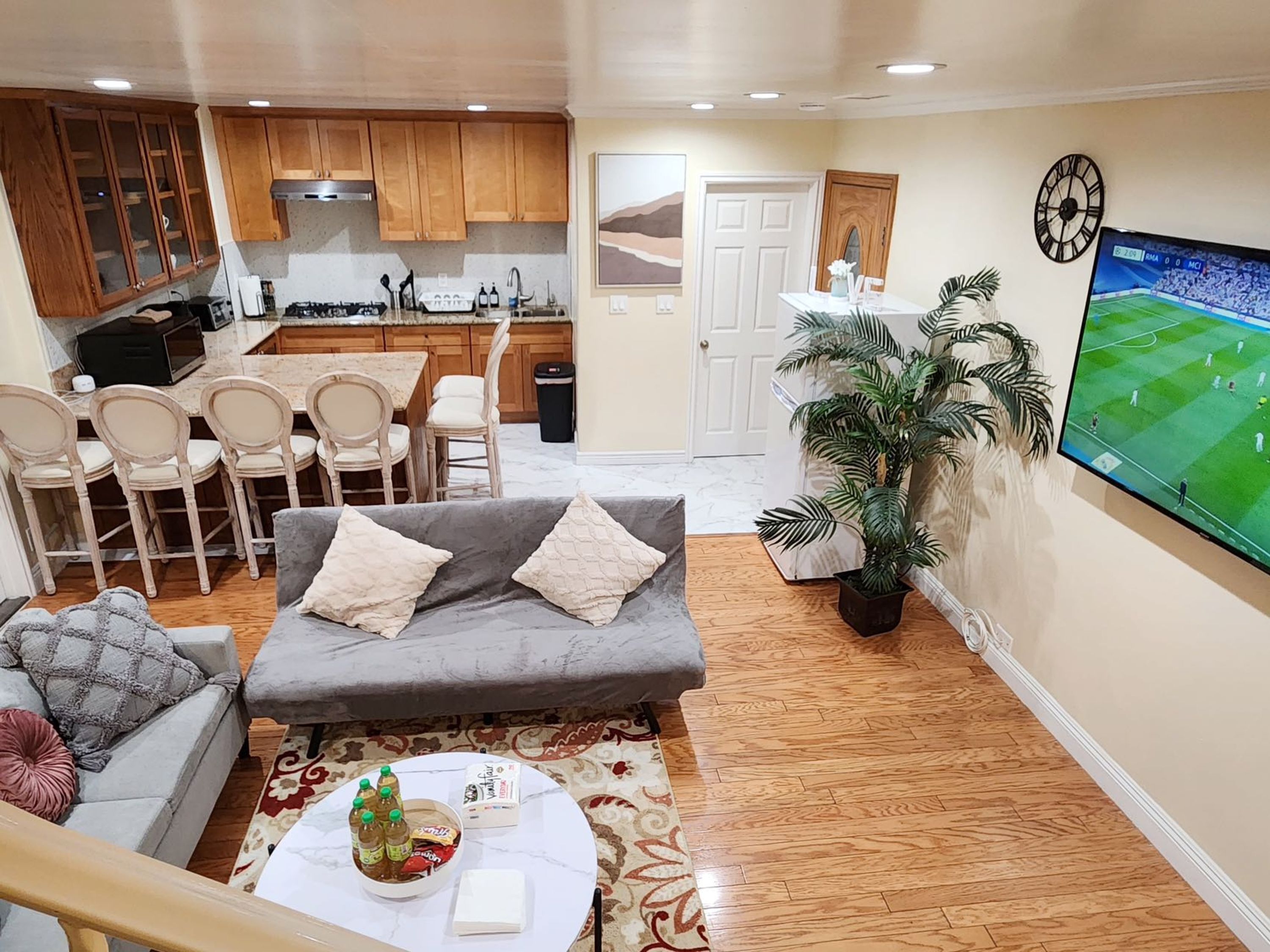 A living room with a grey sofa, a coffee table with snacks, a TV displaying a sports game, and a kitchen with wooden cabinets and a dining area.