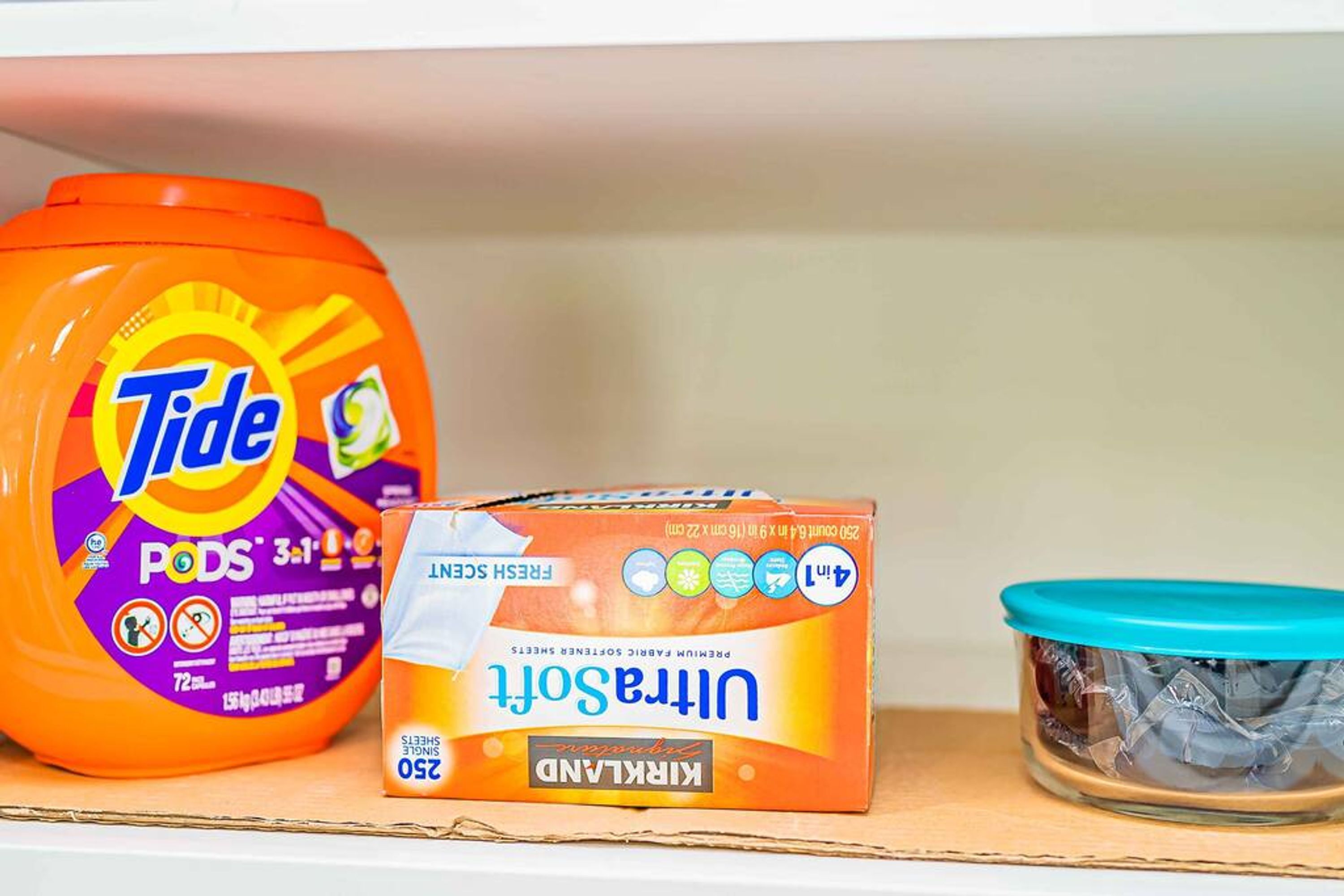 Caixes de Tide PODS, un cartró de detergent Kirkland Ultra Soft i un recipient de vidre amb tapa blava sobre una prestatgeria.