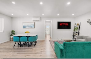 Un soggiorno moderno con un divano verde, un'area pranzo con sedie color petrolio e un televisore a schermo piatto che mostra 'NETFLIX'.